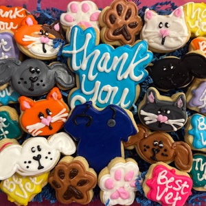 May include: Assortment of decorated sugar cookies with various animal and paw print designs. The cookies are decorated with blue, pink, green, yellow, white, and brown icing. The cookies have text that reads "Thank You", "Best Vet", and "Best Vet".