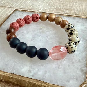 May include: A bracelet made with black lava rock, wood, and dalmatian jasper beads. The bracelet also features a large, faceted, pink crystal bead.
