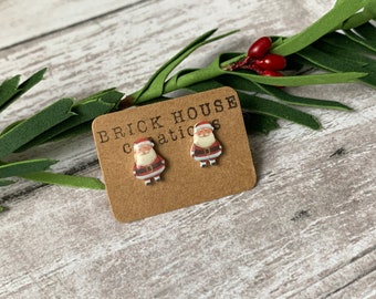 May include: A pair of Santa Claus stud earrings on a brown card with the text "BRICK HOUSE CREATIONS". The earrings feature a red and white Santa design with a black belt and white beard. Greenery and red berries are in the background.