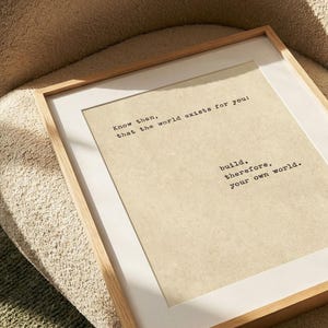 Op de afbeelding: Een ingelijste print met een licht houten frame en witte passe-partout. De print heeft een beige achtergrond met zwarte tekst die luidt: "Weet dan, dat de wereld voor jou bestaat: bouw daarom je eigen wereld."