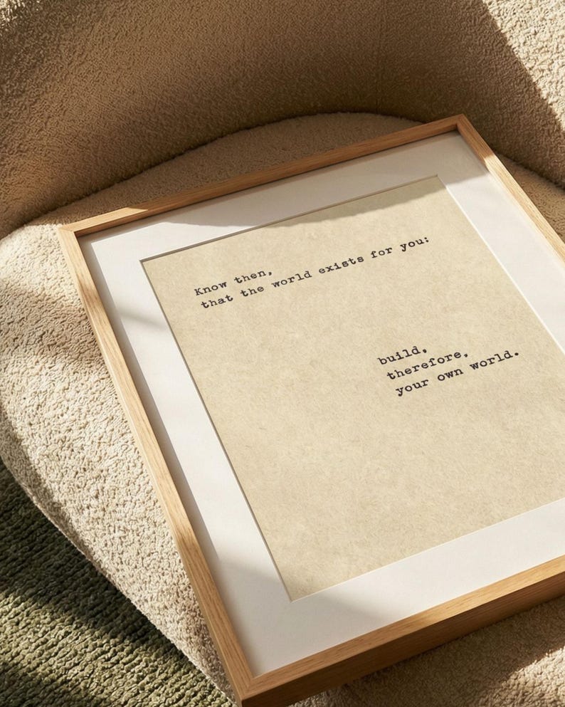 Op de afbeelding: Een ingelijste print met een licht houten frame en witte passe-partout. De print heeft een beige achtergrond met zwarte tekst die luidt: "Weet dan, dat de wereld voor jou bestaat: bouw daarom je eigen wereld."