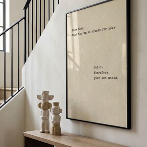 Op de afbeelding: Een ingelijste print met de tekst "Know then, that the world exists for you: build, therefore, your own world." wordt weergegeven boven een houten console. Twee gebeeldhouwde, lichtgekleurde beelden staan op de console.