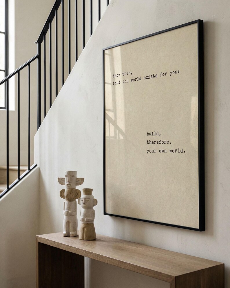 Op de afbeelding: Een ingelijste print met de tekst "Know then, that the world exists for you: build, therefore, your own world." wordt weergegeven boven een houten console. Twee gebeeldhouwde, lichtgekleurde beelden staan op de console.