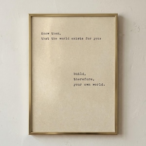 Op de afbeelding: Een ingelijste print met een goudkleurige lijst. De print heeft een beige achtergrond met zwarte tekst die luidt: "Weet dan, dat de wereld voor jou bestaat: bouw daarom je eigen wereld."
