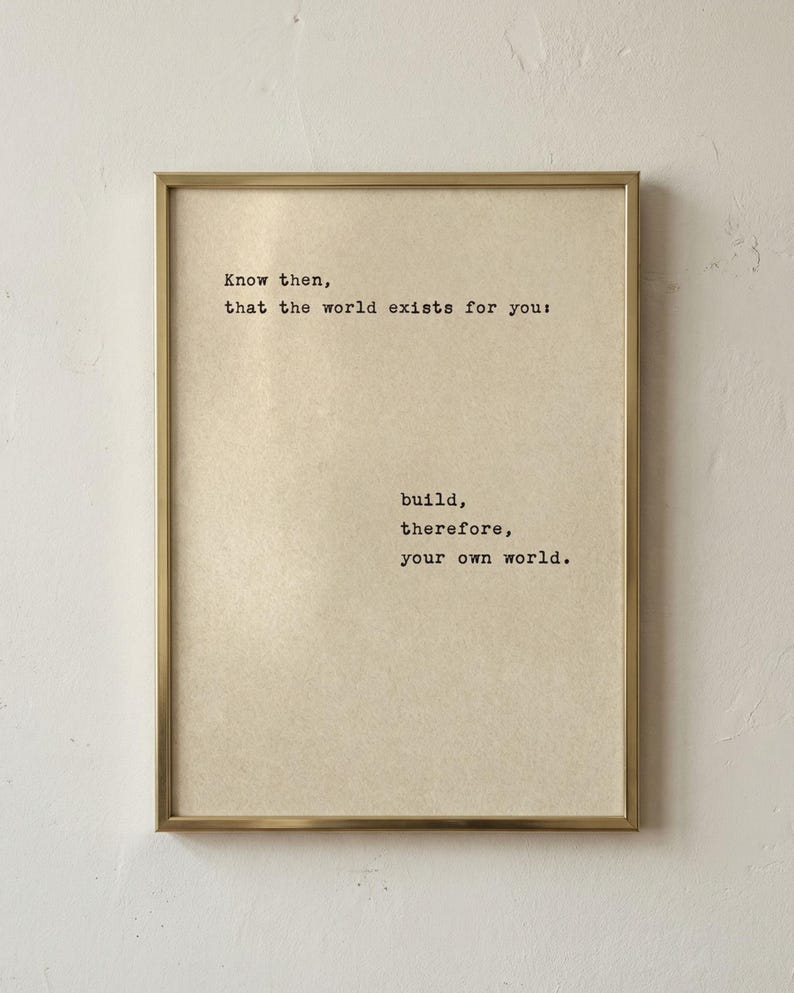 Op de afbeelding: Een ingelijste print met een goudkleurige lijst. De print heeft een beige achtergrond met zwarte tekst die luidt: "Weet dan, dat de wereld voor jou bestaat: bouw daarom je eigen wereld."