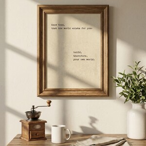Op de afbeelding: Een ingelijste print met de tekst "Weet dan, dat de wereld voor jou bestaat: bouw daarom je eigen wereld." Een houten koffiemolen, witte mok en vaas met groen staan op een houten tafel.