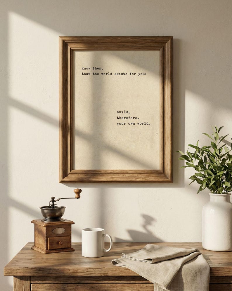 Op de afbeelding: Een ingelijste print met de tekst "Weet dan, dat de wereld voor jou bestaat: bouw daarom je eigen wereld." Een houten koffiemolen, witte mok en vaas met groen staan op een houten tafel.