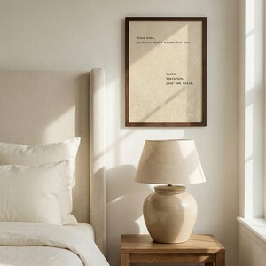 Op de afbeelding: Slaapkamerinterieur met een ingelijste print met de tekst "Know then, that the world exists for you: build, therefore, your own world." Een beige lamp staat op een houten nachtkastje naast een bed met witte lakens.