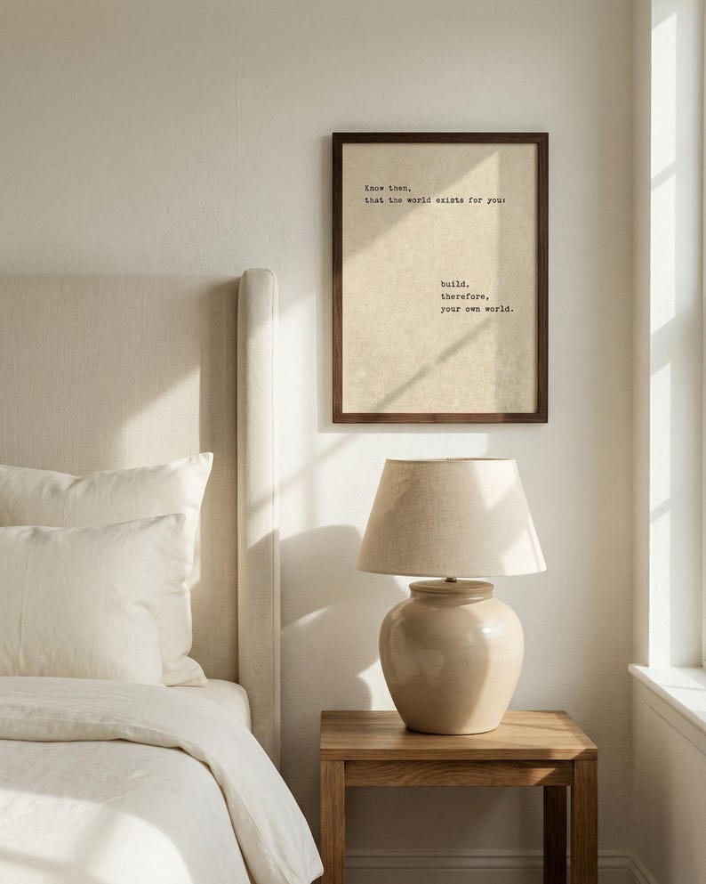 Op de afbeelding: Slaapkamerinterieur met een ingelijste print met de tekst "Know then, that the world exists for you: build, therefore, your own world." Een beige lamp staat op een houten nachtkastje naast een bed met witte lakens.