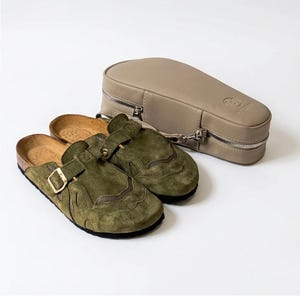 Könnte beinhalten: Ein Paar olivgrüne Wildleder-Clogs mit seitlichem Schnallenriemen und strukturierter Sohle, begleitet von einem passenden beigen, strukturierten Tragekoffer mit silbernem Reißverschluss und einem geprägten Logo auf der Oberseite.
