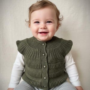 May include: A smiling baby wearing an olive green knitted vest with three button closures. The vest is layered over a white long-sleeved shirt and paired with gray pants. The baby has light brown hair.