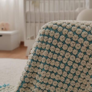 May include: A cream-colored, textured baby blanket with a floral pattern and a turquoise border. The blanket is made of soft material and is likely handmade. The blanket is displayed in a nursery setting.
