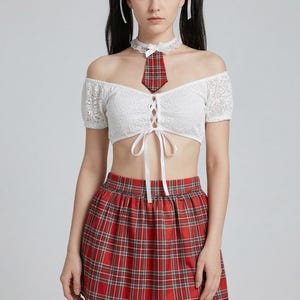 Op de afbeelding: Een witte kanten off-the-shoulder crop top en een rode geruite minirok. De outfit bevat een bijpassende rode geruite stropdas en choker. Het model heeft zwart haar in staartjes. De rok is een klassieke schooluniformstijl.
