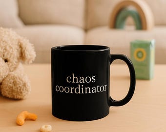 以下が含まれることがあります： 白い文字で「chaos coordinator」と書かれた黒いセラミックマグカップ。 マグカップには湾曲したハンドルがあり、明るい色の木製の表面に置かれています。 表面には、ぬいぐるみ、軽食、装飾品などのアイテムがあります。