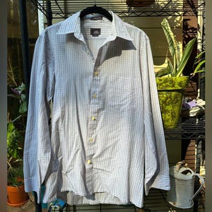 May include: A long-sleeved shirt with a light blue and white checked pattern. The shirt features a classic collar, a chest pocket, and gold-coloured buttons. The brand name "John Henry" is visible on the label.