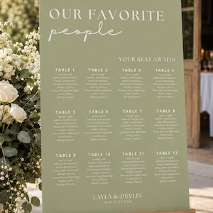 May include: A sage green rectangular wedding seating chart sign on a wooden easel. The sign features white text reading OUR FAVORITE people, YOUR SEAT AWAITS, tables 1 through 12 with guest names, and the names LAYLA & DEVLIN with AUGUST 23, 2026.