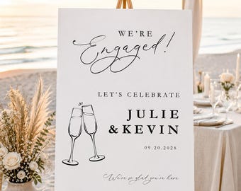 May include: A white rectangular wedding sign on a wooden easel featuring the black text We're Engaged!, Let's Celebrate, Julie & Kevin, the date 09.20.2028, and We're so glad you're here, with a black line drawing of two clinking champagne flutes.