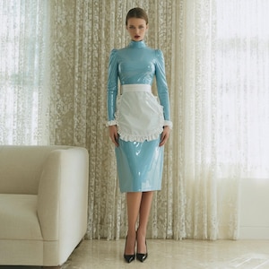 May include: A light blue, high-neck dress with long sleeves and a matching skirt. The dress is paired with a white apron with ruffled edges. The model is wearing black heels. The background includes a white sofa and patterned curtains.