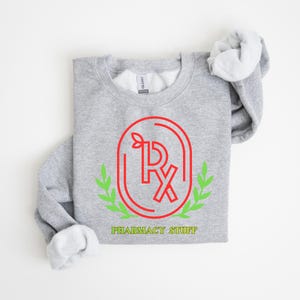 May include: A heather gray sweatshirt with a red and green pharmacy logo. The logo features the letters "Rx" inside an oval frame, with green leaves and the words "PHARMACY STUFF" in green below.