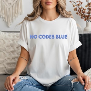 May include: White t-shirt with the text "NO CODES BLUE" in blue, sans-serif font. The shirt has short sleeves and a relaxed fit. The model is wearing blue jeans.