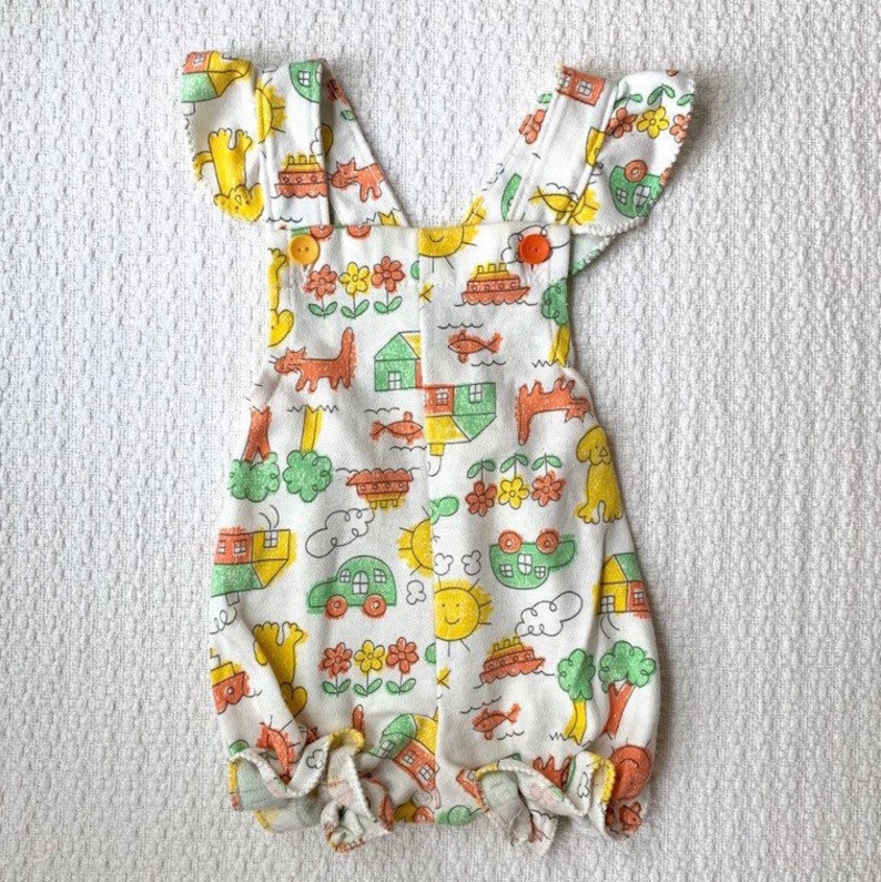 May include: White baby overalls with a colorful print of houses, cars, fish, and suns. The overalls have ruffle trim on the straps and legs.