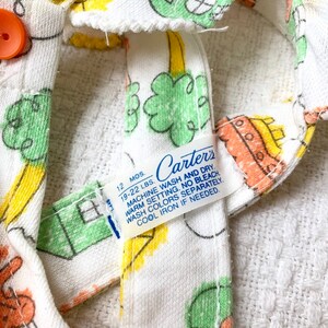 May include: White baby onesie with a green, yellow, and orange print of a house, a tree, and a rocket. The onesie has a blue Carter's label with the text "12 MOS. 19-22 LBS. MACHINE WASH AND DRY. WARM SETTING. NO BLEACH WASH COLORS SEPARATELY. COOL IRON IF NEEDED."
