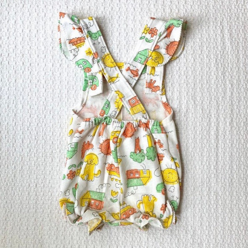 May include: White baby romper with a yellow, green, and orange print of houses, suns, and animals. The romper has a criss-cross strap design in the back.