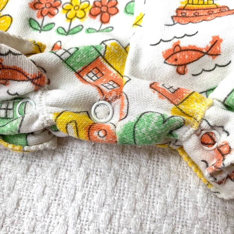 May include: Close-up of the white fabric cuff of a baby onesie with a colorful cartoon print of houses, flowers, and a fish. The cuff has two white plastic snaps.