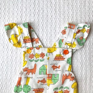 May include: White overalls with a playful design featuring houses, boats, cats, and fish. The straps have a ruffled edge and the overalls have two yellow buttons.