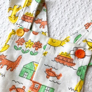 May include: White cotton overalls with a playful design featuring yellow suns, orange fish, red houses, and green trees. The overalls have two yellow buttons and orange straps.