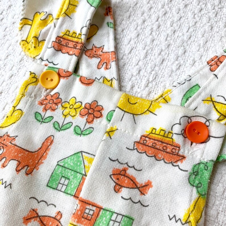 May include: White cotton overalls with a playful design featuring yellow suns, orange fish, red houses, and green trees. The overalls have two yellow buttons and orange straps.