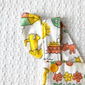May include: White cotton overalls with a yellow, orange, and green print of animals, a boat, and a sun. The overalls have a yellow button.