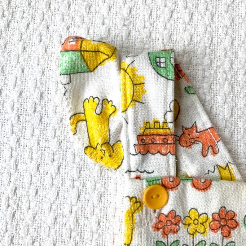 May include: White cotton overalls with a yellow, orange, and green print of animals, a boat, and a sun. The overalls have a yellow button.