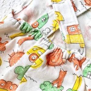 May include: White cotton fabric with a yellow, orange, and green print of houses, suns, and trees. The fabric has a gathered elastic band at the top.