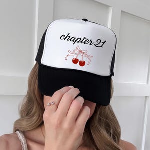May include: A black and white trucker hat featuring the words "chapter 21" in black script, with a red cherry illustration. The hat is worn by a person. The hat has a mesh back.