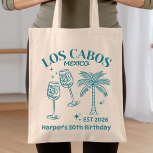 May include: A cream tote bag featuring teal text and illustrations. The design includes "Los Cabos Mexico", two wine glasses, a palm tree, and the text "EST 2026" and "Harper's 30th Birthday".