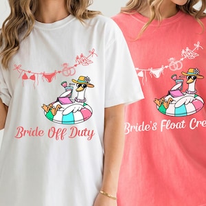 May include: Two t-shirts, one white and one coral, feature a cartoon goose in a floatie, wearing a hat and sunglasses, with a drink. The white shirt says "Bride Off Duty" and the coral shirt says "Bride's Float Crew". Both shirts have a bikini and wedding ring design.