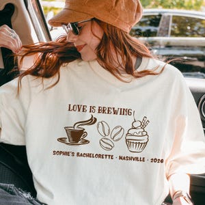 Può includere: T-shirt color crema con la scritta "LOVE IS BREWING" e grafica a tema caffè. Il design include una tazza di caffè, chicchi di caffè e un cupcake. Il testo "SOPHIE'S BACHELORETTE - NASHVILLE - 2026" è stampato anche sulla maglietta.