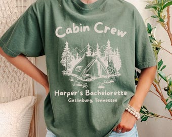 May include: Green t-shirt with the text "Cabin Crew" above a white graphic of a tent, trees, and mountains. Below the graphic, the text reads "Harper's Bachelorette Gatlinburg, Tennessee."