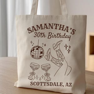 May include: Beige tote bag with a brown graphic design. The design includes text that reads "SAMANTHA'S 30th Birthday" and "SCOTTSDALE, AZ". The graphic also features a disco ball, cowboy boots, and two wine glasses.