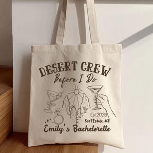 May include: Cream-colored tote bag with the text "Desert Crew Before I Do" and "Emily's Bachelorette". The bag features a desert-themed graphic with cocktails, a ring, cacti, and the text "Est. 2026 Scottsdale, AZ".