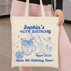 May include: Cream-colored tote bag with a blue illustration. The design includes text that reads "Sophia's 40th Birthday" and "Aprés Ski Birthday Crew!" with images of a mug, mountains, snowflakes, and winter accessories.