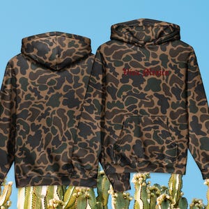 Puede incluir: Dos sudaderas con capucha con un estampado de camuflaje marrón, verde y negro. La sudadera de la derecha tiene el texto "Pine Bluffs" en rojo en el pecho. Las sudaderas tienen un bolsillo delantero y una capucha.