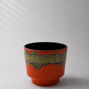 Puede incluir: Una maceta cilíndrica de cerámica que presenta un color base naranja vibrante con una banda texturizada de color verde musgo que rodea la parte superior. El interior del recipiente tiene un acabado de esmalte negro sólido y el exterior es liso y brillante.