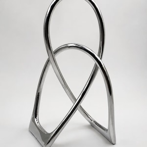 Puede incluir: Una escultura de metal plateado pulido que presenta dos bucles continuos entrelazados que forman una figura abstracta tridimensional. La superficie lisa y reflectante se curva hacia una base estable con cuatro puntos de apoyo.