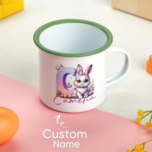 Könnte beinhalten: Weißer Emaille-Becher mit grünem Rand und Henkel. Der Becher zeigt ein Cartoon-Kaninchen mit rosa Schleife, den Buchstaben "C" und den Namen "Camelia". Darunter steht "Custom Name".