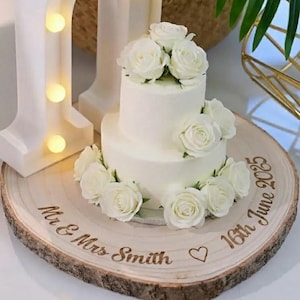 Puede incluir: Una tarta de boda blanca de dos pisos decorada con rosas blancas, presentada sobre una rodaja de madera. La rodaja está grabada con los nombres "Mr & Mrs Smith", un corazón y la fecha "16 de junio de 2025".