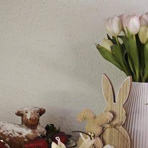 Può includere: Natura morta pasquale con una torta a forma di agnello, fragole e due candele a forma di coniglietto su una fetta di legno. Un coniglietto di legno e un mazzo di tulipani bianchi e rosa completano la scena.