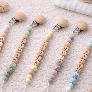 May include: Four personalized baby pacifier clips with wooden beads spelling out names: MILA, ETHAN, LIVER, LUCY, and AYDEN. Each clip has a wooden clip, a variety of colorful beads, and a silicone pacifier holder.