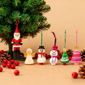 Op de afbeelding: Een verzameling kerstboomversieringen, waaronder een kerstman, een engel, sneeuwmannen en kerstbomen. De ornamenten zijn in verschillende kleuren, zoals rood, wit, groen en roze, en hangen voor een kleine kerstboom.